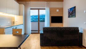 A modern kitchen with a dining table and a sofa, a TV mounted on the wall, and a large window with a mountain view.