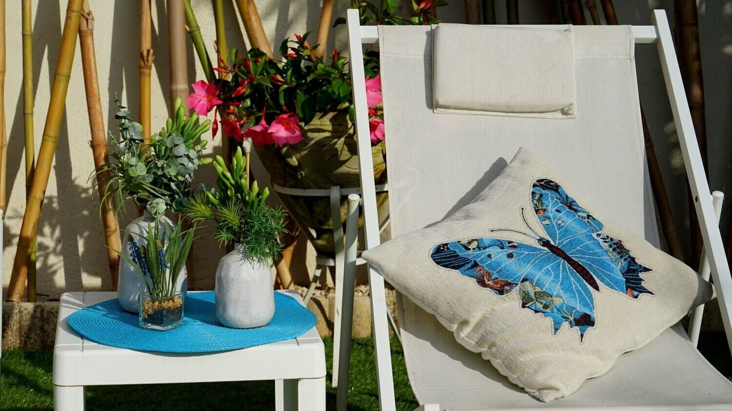 A white lounge chair with a cushion featuring a blue butterfly design, a small table with a blue mat and two potted plants, and a bamboo backdrop.