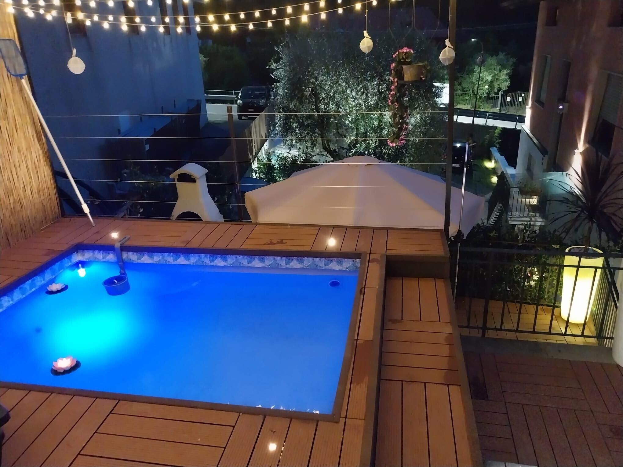 A rooftop pool with a wooden deck, string lights, and a view of the cityscape.