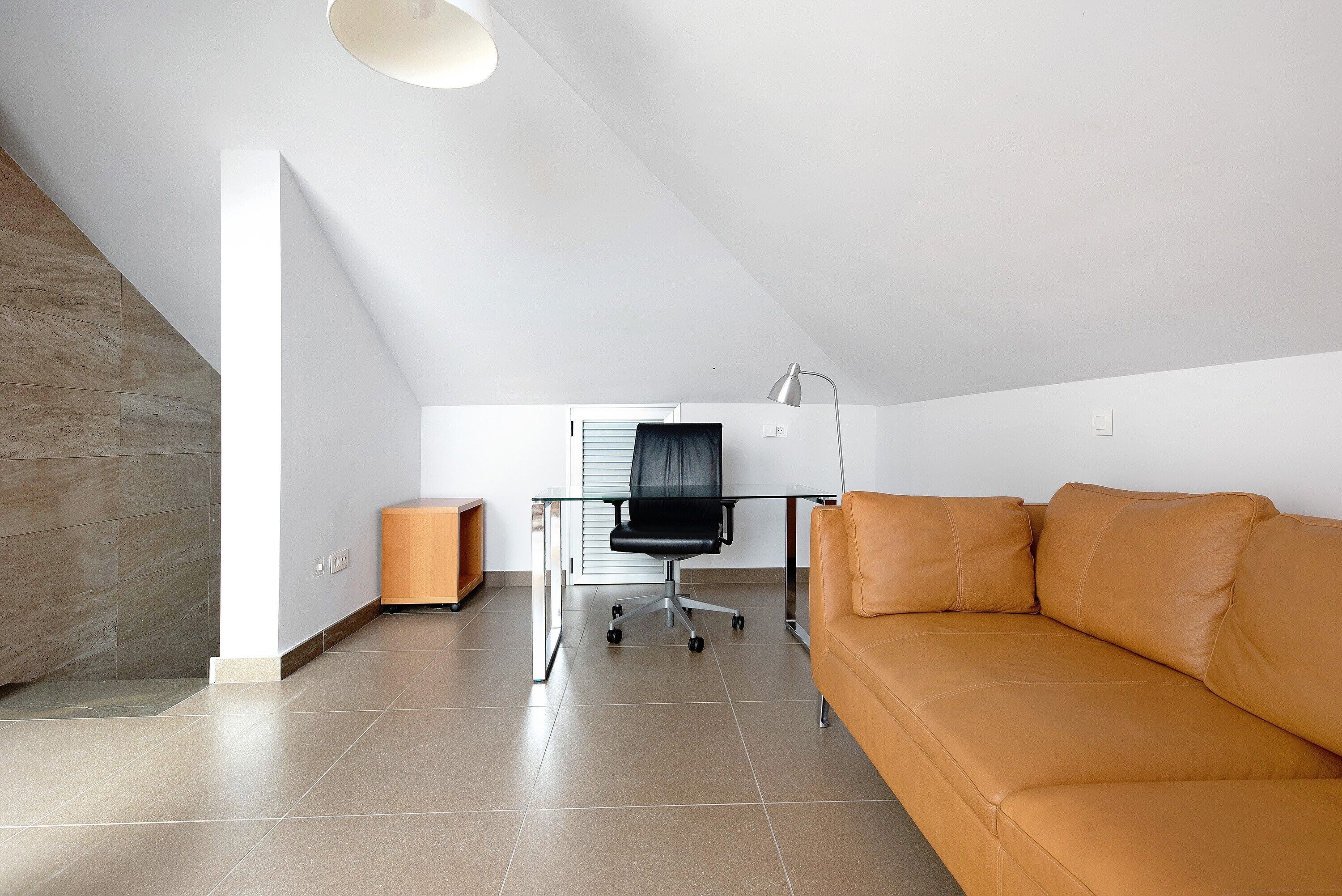 Una habitación moderna con techo inclinado, un escritorio de vidrio, una silla de oficina negra, un sofá de cuero marrón y suelo de madera.