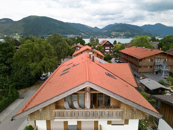 A wooden chalet with a sloped roof, a balcony with seating, and a view of a lake and mountains in the background.