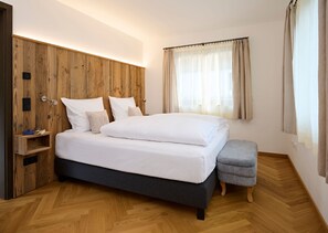 A modern bedroom with a wooden headboard, a bed with white linens, a bedside table with books, and a window with curtains. - Ferienappartement Trachtenhut - Alpenseensucht Natur-ferienappartements (Bad Wiessee)