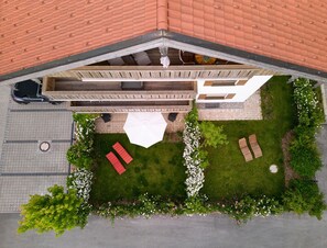 Ovenfra ses en bolig med skråt tag, en belagt indkørsel, en græsplæne med en hvid parasol og en træterrasse.
