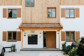 A building with a wooden facade, multiple windows with shutters, a door, and a small garden area with plants. - Ferienappartement Trachtenhut - Alpenseensucht Natur-ferienappartements (Bad Wiessee)