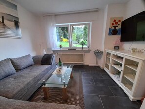 A living room with a sofa, a glass coffee table, a white cabinet, a TV, and a window with a view of greenery. - Fewo Ida-weinbergblick (Traben-Trarbach)