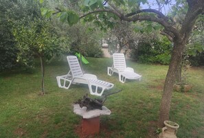 Deux chaises longues en plastique blanc, placées sous un arbre dans un espace extérieur verdoyant.