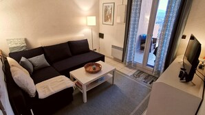 Un salon avec un canapé foncé, une table basse blanche et un téléviseur. Il y a une fenêtre avec des rideaux, un radiateur et un tableau encadré au mur.