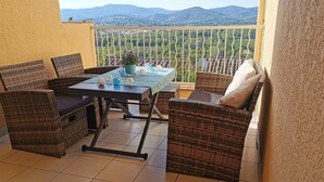 Un balcon agrémenté de meubles en osier, d’une table en verre et offrant une vue sur des collines lointaines.