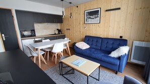A modern living room with a blue sofa, a dining table with chairs, and a kitchen area with dark cabinets and a coffee machine. - Appartement 2 Pieces Pied DES Pistes Proche Centre Balnéo et Randonnées (Aragnouet)