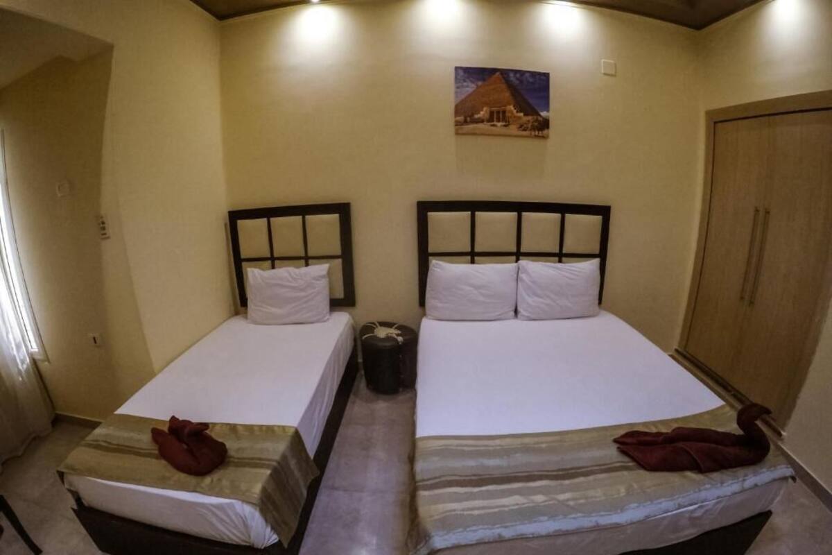 A room with two beds, each with a headboard and pillows. There is a small trash bin between the beds.