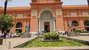 Un grand bâtiment historique doté d’une entrée centrale en arc de cercle, entouré d’un jardin aménagé avec un élément décoratif aquatique.