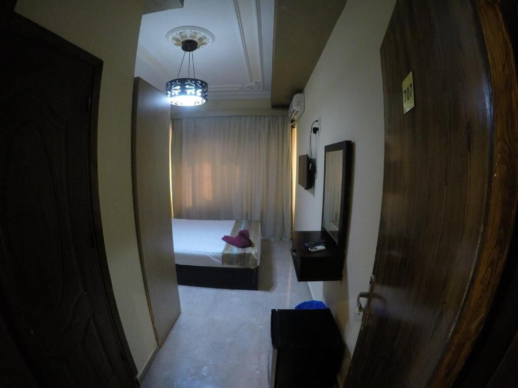 Un couloir étroit mène à une chambre à coucher qui contient un lit, une table de chevet, un miroir et un luminaire fixé au plafond.