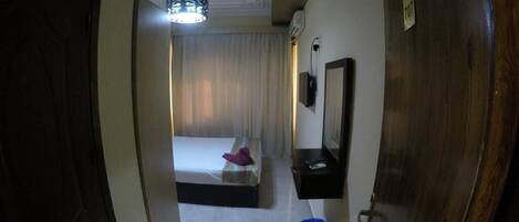 Un couloir étroit mène à une chambre à coucher qui contient un lit, une table de chevet, un miroir et un luminaire fixé au plafond.