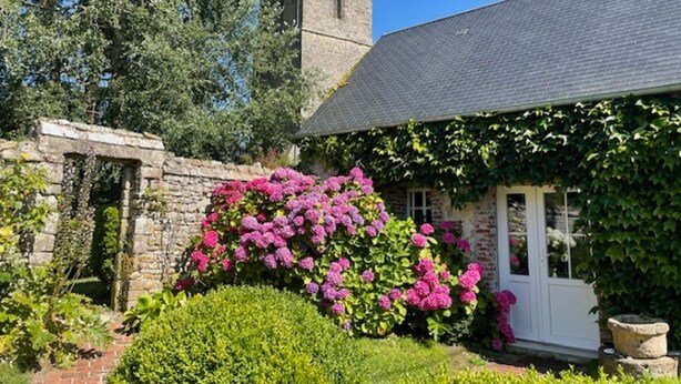 Een stenen gebouw met een toren, omringd door weelderige begroeiing en felroze bloemen.