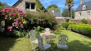 Een stenen huis met een tuin met roze bloemen en een klein terras met metalen stoelen en een tafel.