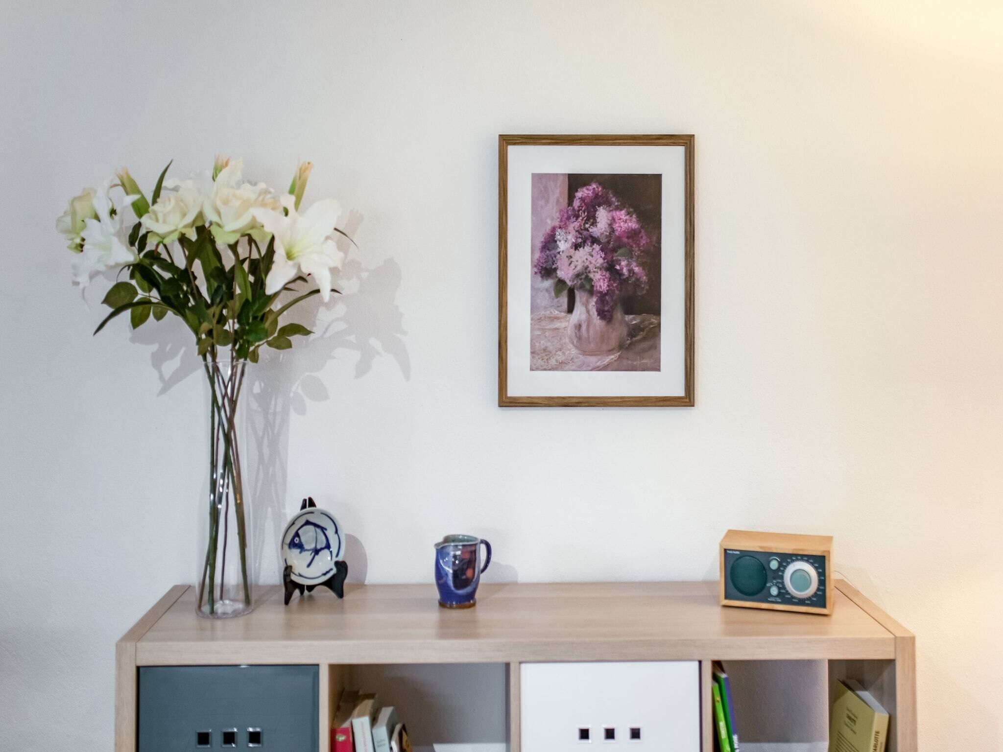 Et træskrivebord med en vase fyldt med hvide blomster, et indrammet billede, et ur, en kop og en radio.