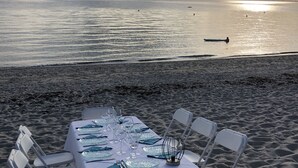 Een tafel gedekt voor zes personen op een zanderig strand, met uitzicht op de oceaan en de ondergaande zon.