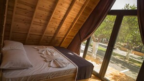 Un dormitorio con techo inclinado, una cama con colchón blanco y ropa de cama marrón, y un ventanal con vistas a árboles y una valla.