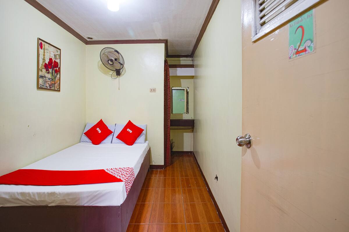 A hotel room with a bed, a fan, a door, and a wall with a picture.