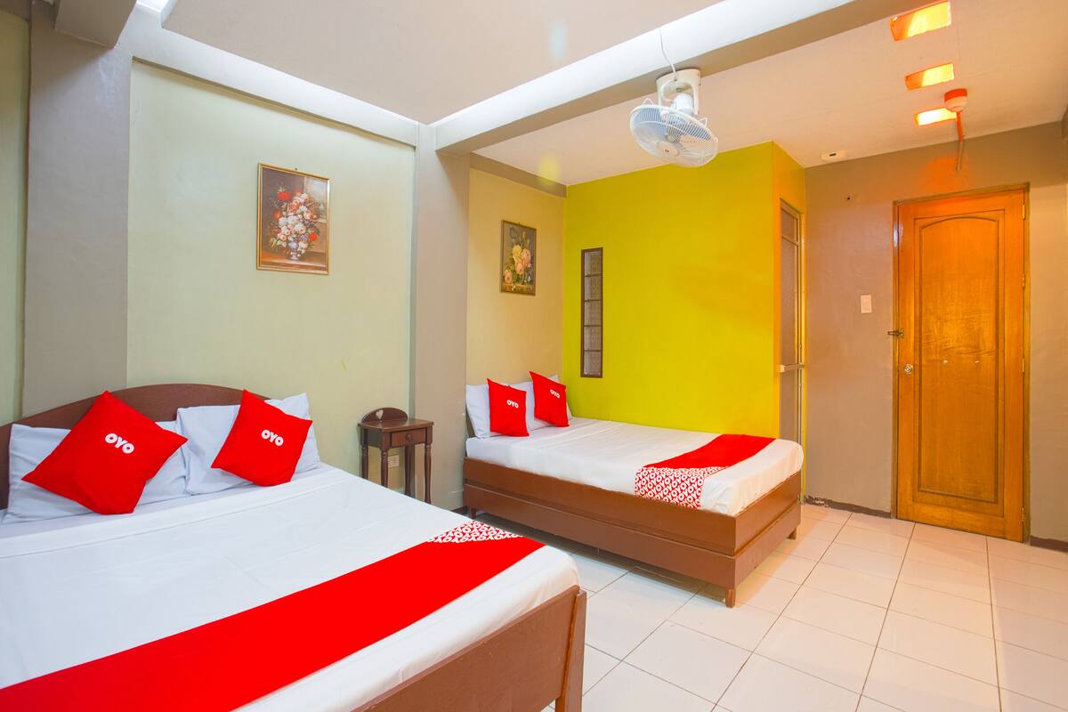 A hotel room with two beds, each with red and white bedding, a small bedside table, a nightstand, and a ceiling fan.