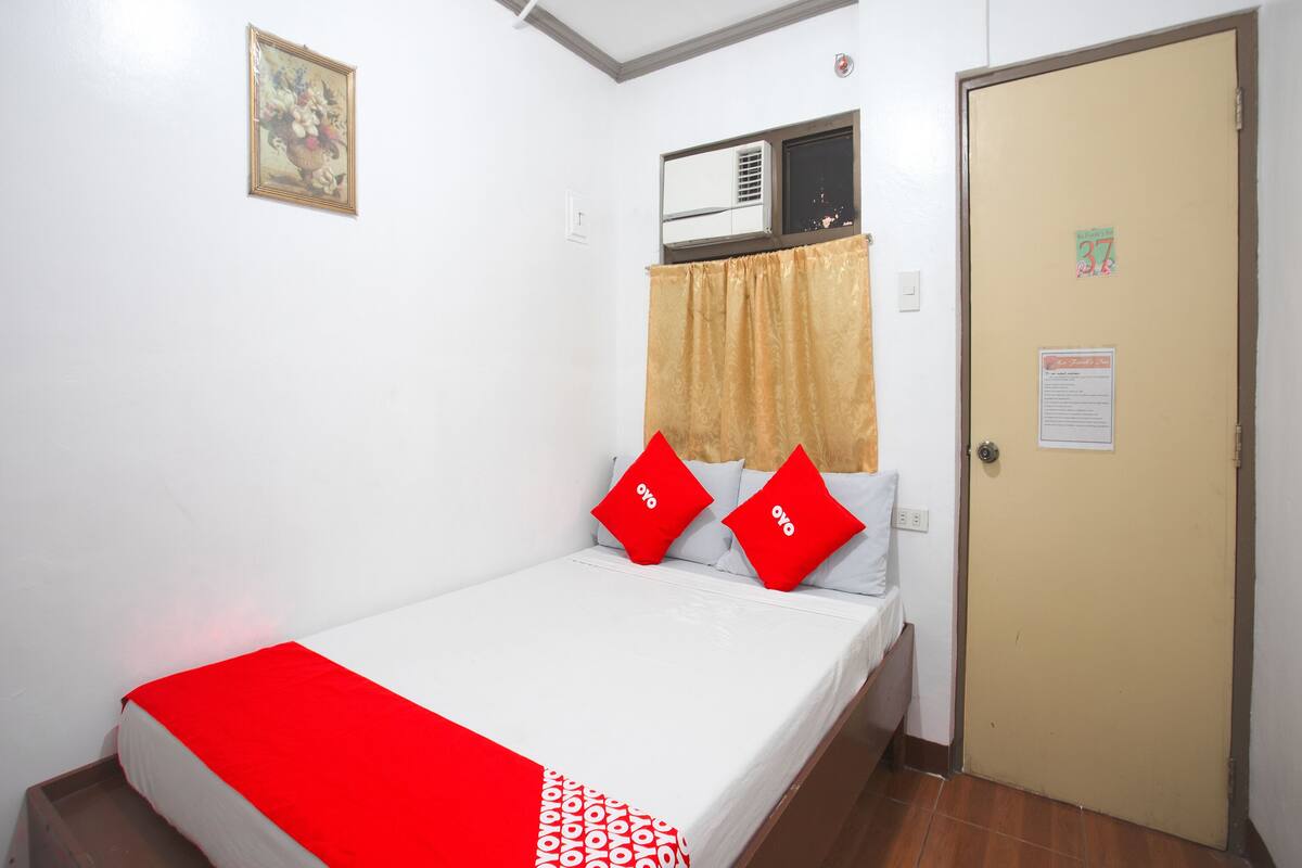A single bed with a red and white bedspread, two red pillows, a beige curtain, and a door with a sign.
