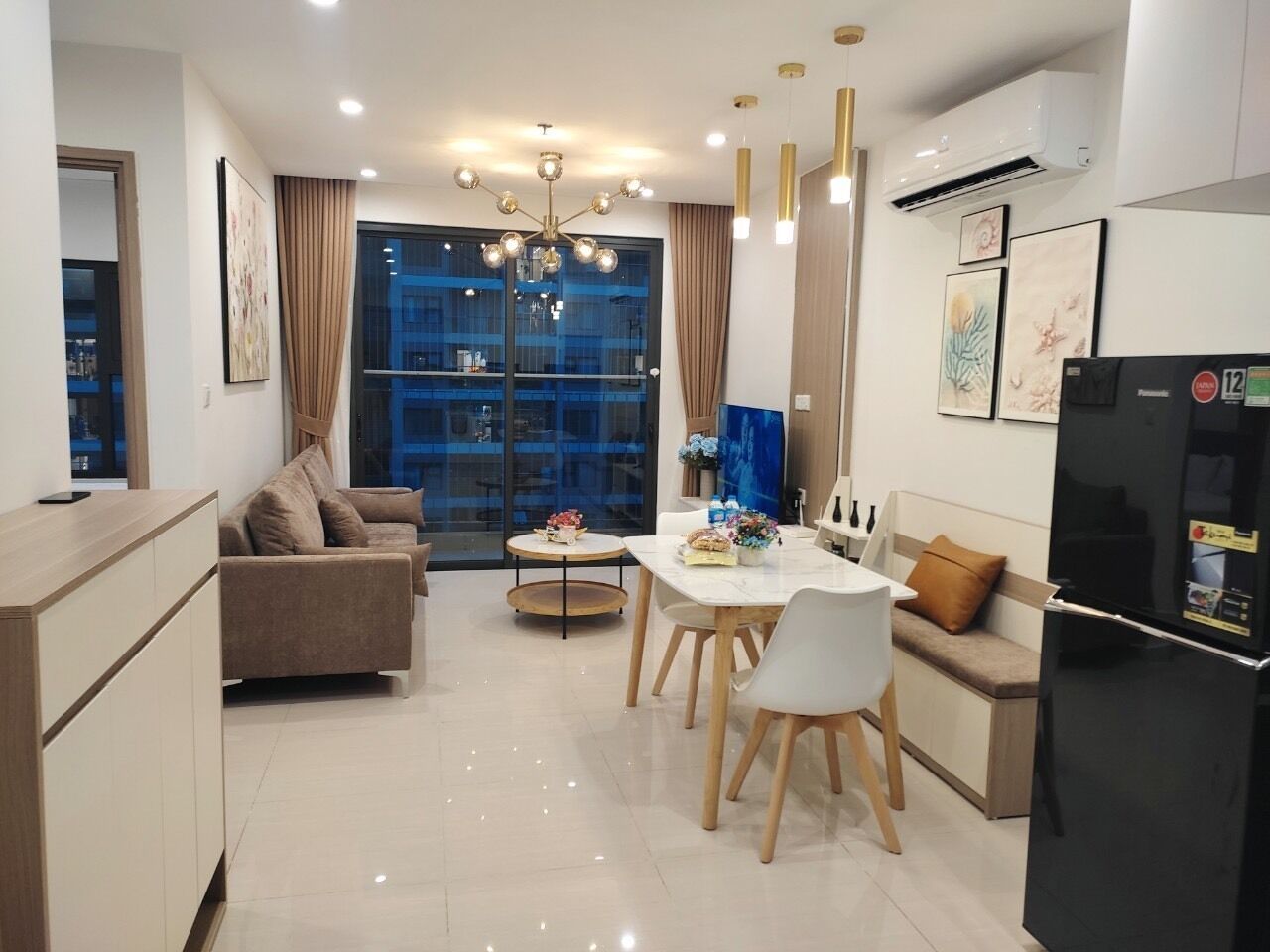 A modern living room with a sofa, dining table, and chairs, a kitchen area with a refrigerator, and a large window with curtains.
