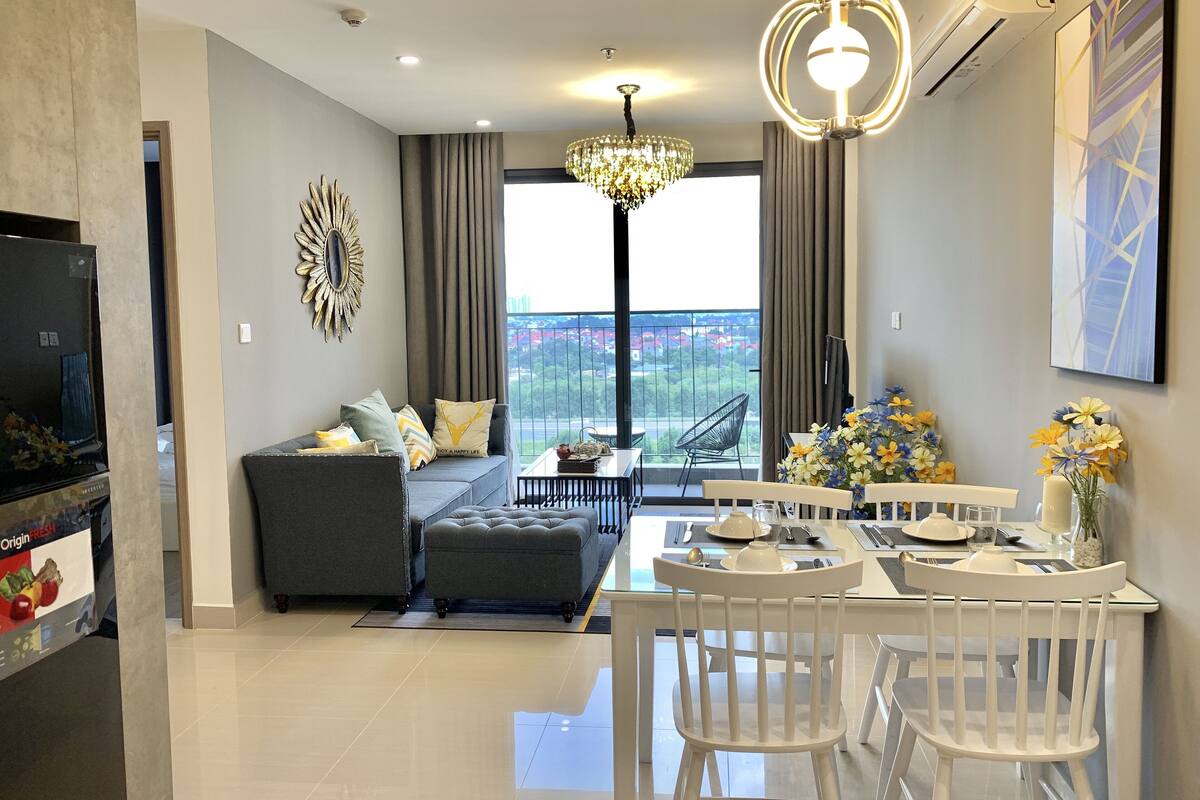 A modern living room with a grey sofa, a dining table with white chairs, a chandelier, and a large window with a view of greenery.