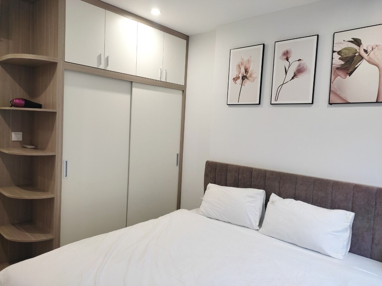 A bedroom with a bed, white bedding, a wooden wardrobe, and three framed floral pictures on the wall.