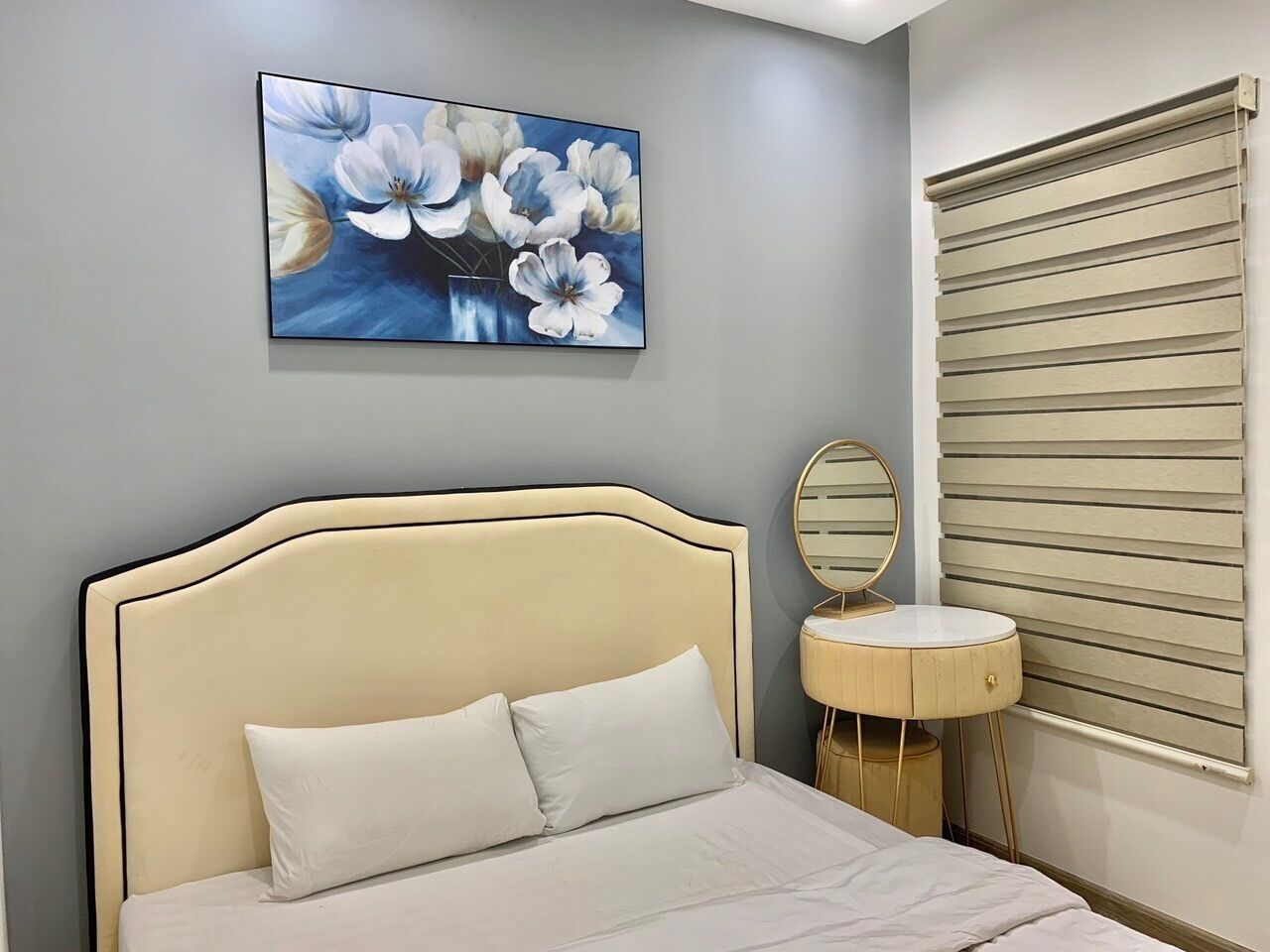 A bedroom with a bed, a bedside table, a round mirror, and a window with blinds.