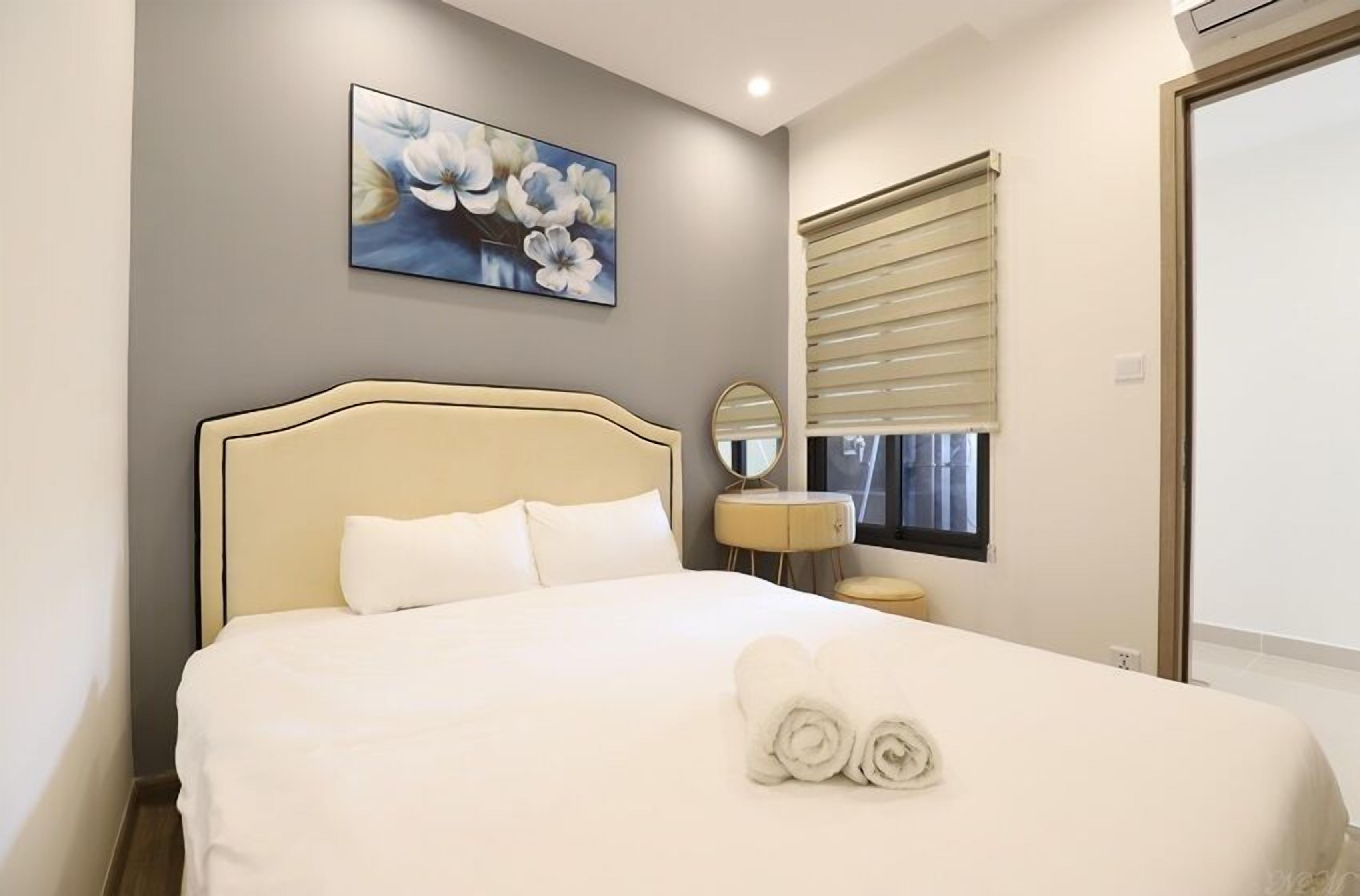 A neatly made bed with white linens and rolled towels, a round mirror, and a floral painting on the wall.