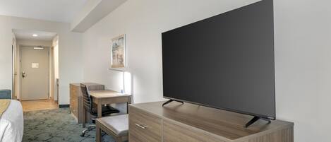Habitación de hotel con cama, escritorio con silla, televisor sobre un mueble de madera y un cuadro en la pared.