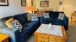 A living room with a dark blue sofa set, patterned throw pillows, a wooden coffee table with a lace table runner, and a framed picture on the wall. - Cozy vacation condo sleeps 6. #201 (Lincoln)