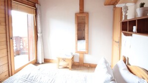 A cozy room with a bed, a small wooden table, a mirror, a shelf with decorative items, and a window with curtains. - Spacious 4-room apartment well equipped with Wi-Fi and parking, at the foot of the ski bus (Les Carroz d'Arâches)