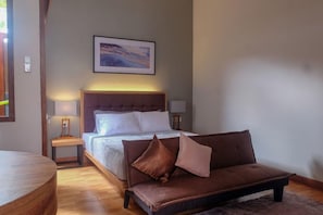 Un dormitorio con una cama, un sofá marrón, una mesita de noche con una lámpara y un cuadro en la pared.