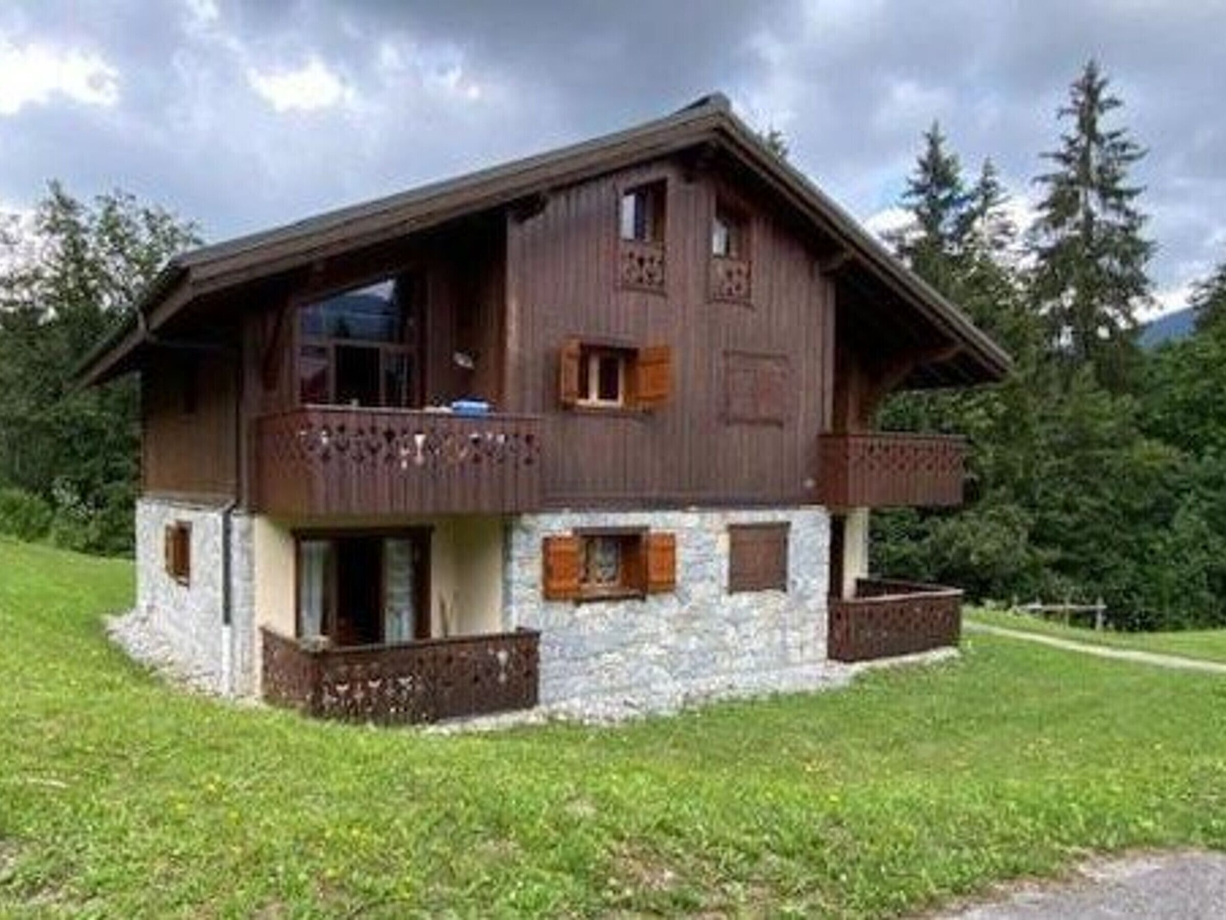A two-story wooden chalet with a stone foundation, a balcony on the upper floor, and a surrounding grassy area.