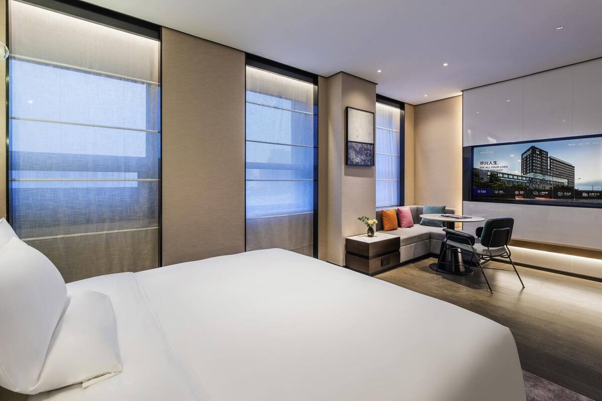 A modern hotel room with a large bed, a small seating area, a TV mounted on the wall, and large windows.