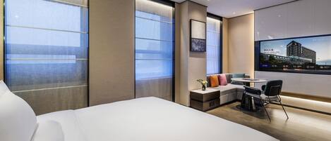A modern hotel room with a large bed, a small seating area, a TV mounted on the wall, and large windows.
