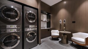 A modern laundry room with four stacked washers and dryers, a small seating area with two chairs, and a round table. - Novotel Tianjin Drum Tower (Tianjin)