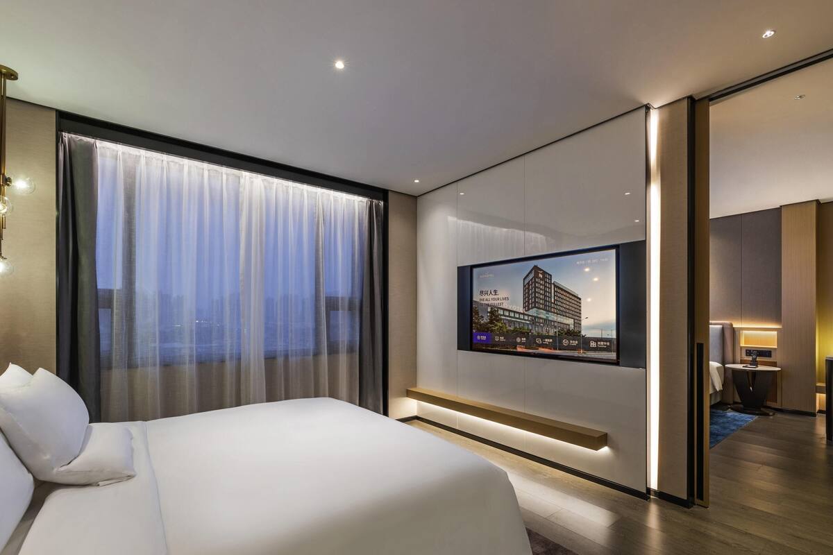A modern hotel room with a large bed, a TV mounted on the wall, and a view of the cityscape through the window.