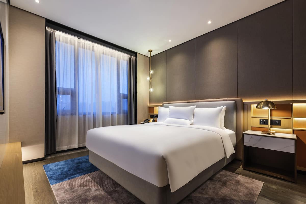 A modern hotel room with a large bed, a bedside table with a lamp, and a view of the city through sheer curtains.