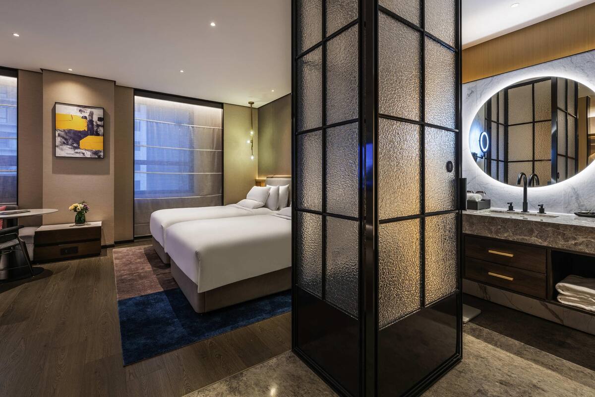 A modern hotel room with a large bed, a separate bathroom with a round mirror, and a view of the city through large windows.