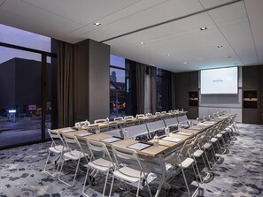 A conference room with a long table, white chairs, a projector screen, and a view of the cityscape through large windows.