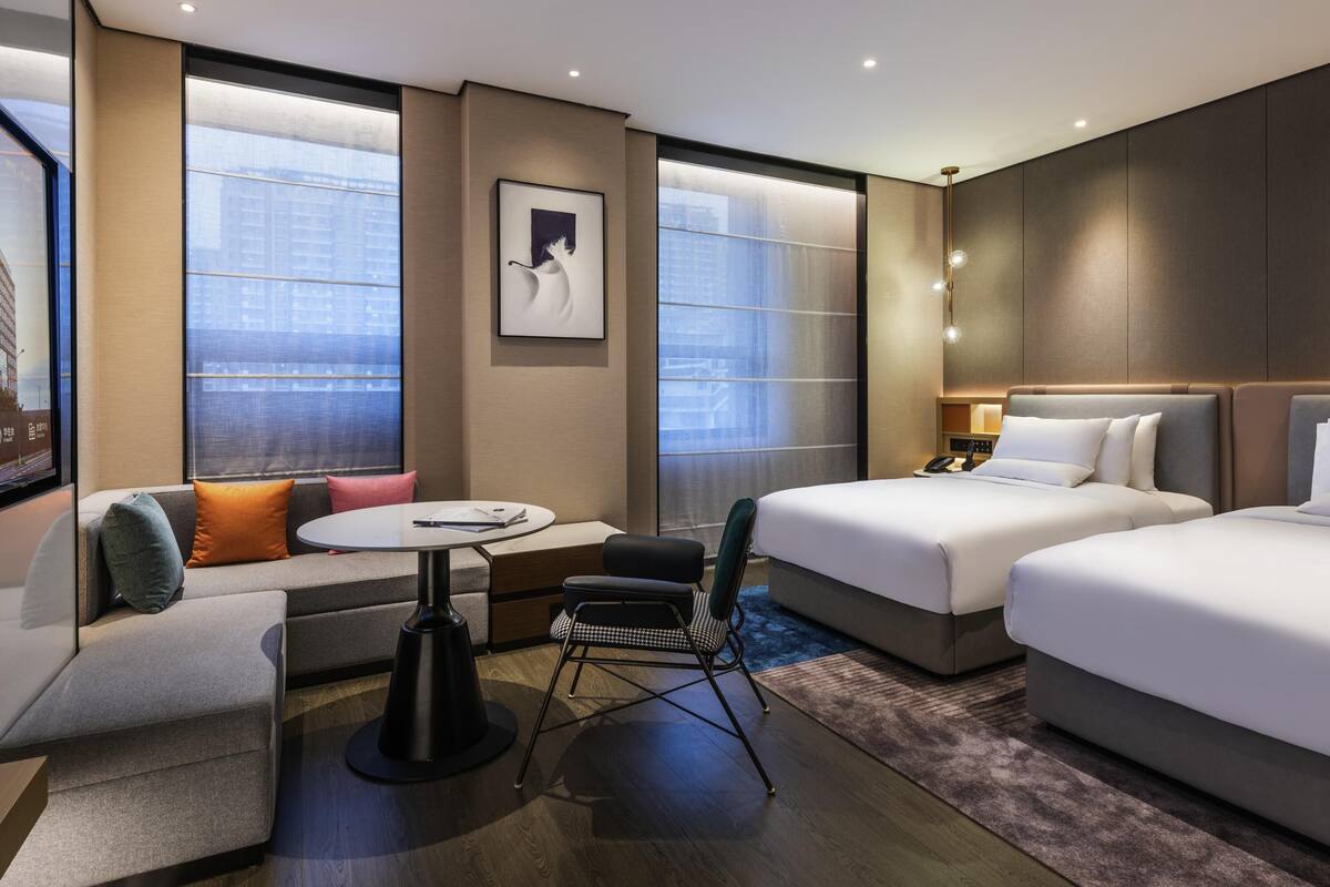 A modern hotel room with a sofa, two beds, a small table, and a chair.