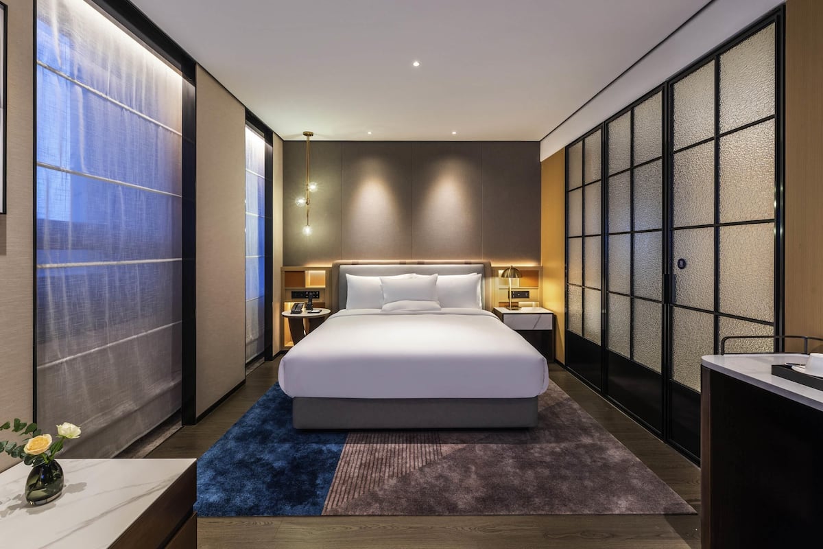 A modern hotel room with a large bed, bedside tables, a nightstand, a desk, and a view of the cityscape.