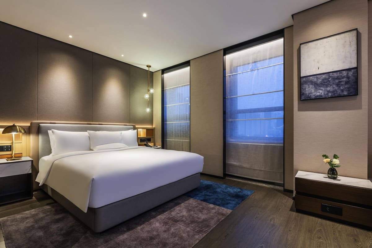 A modern hotel room with a large bed, bedside tables, a nightstand, a desk, a chair, a window with a view, and a painting on the wall.