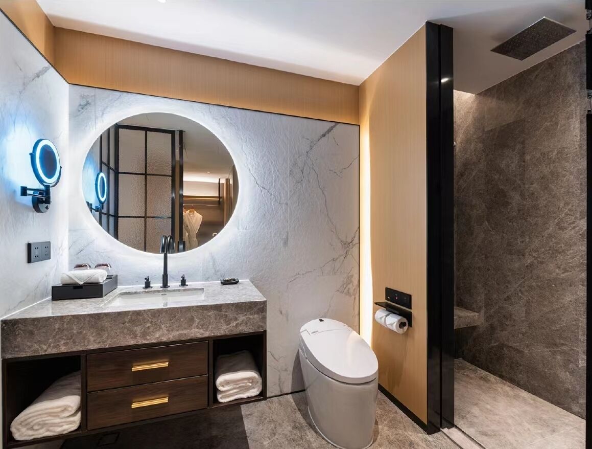 A modern bathroom with a marble vanity, a round mirror, a toilet, and a shower area.