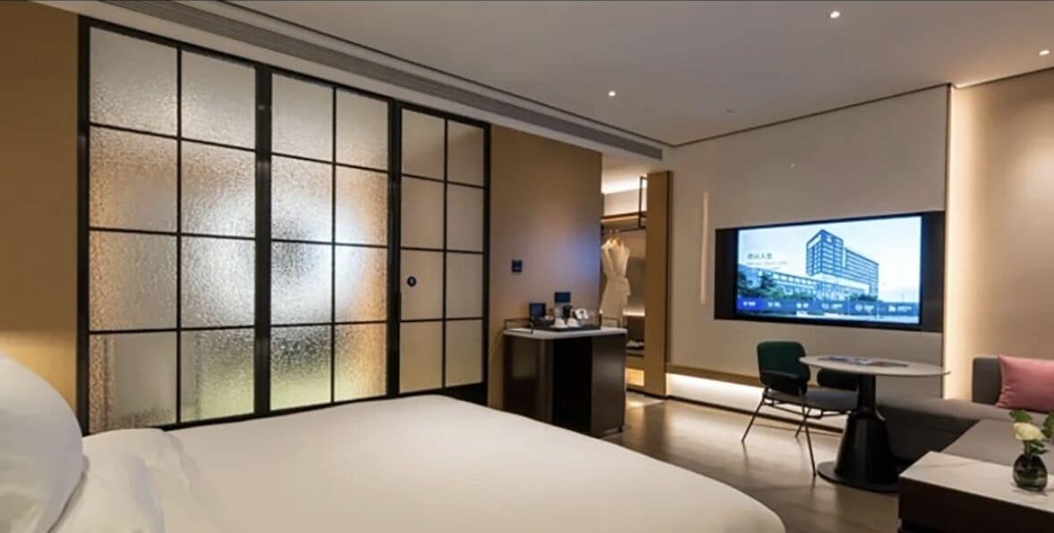 A hotel room with a bed, a desk with a chair, a television mounted on the wall, and a sliding door with frosted glass panels.