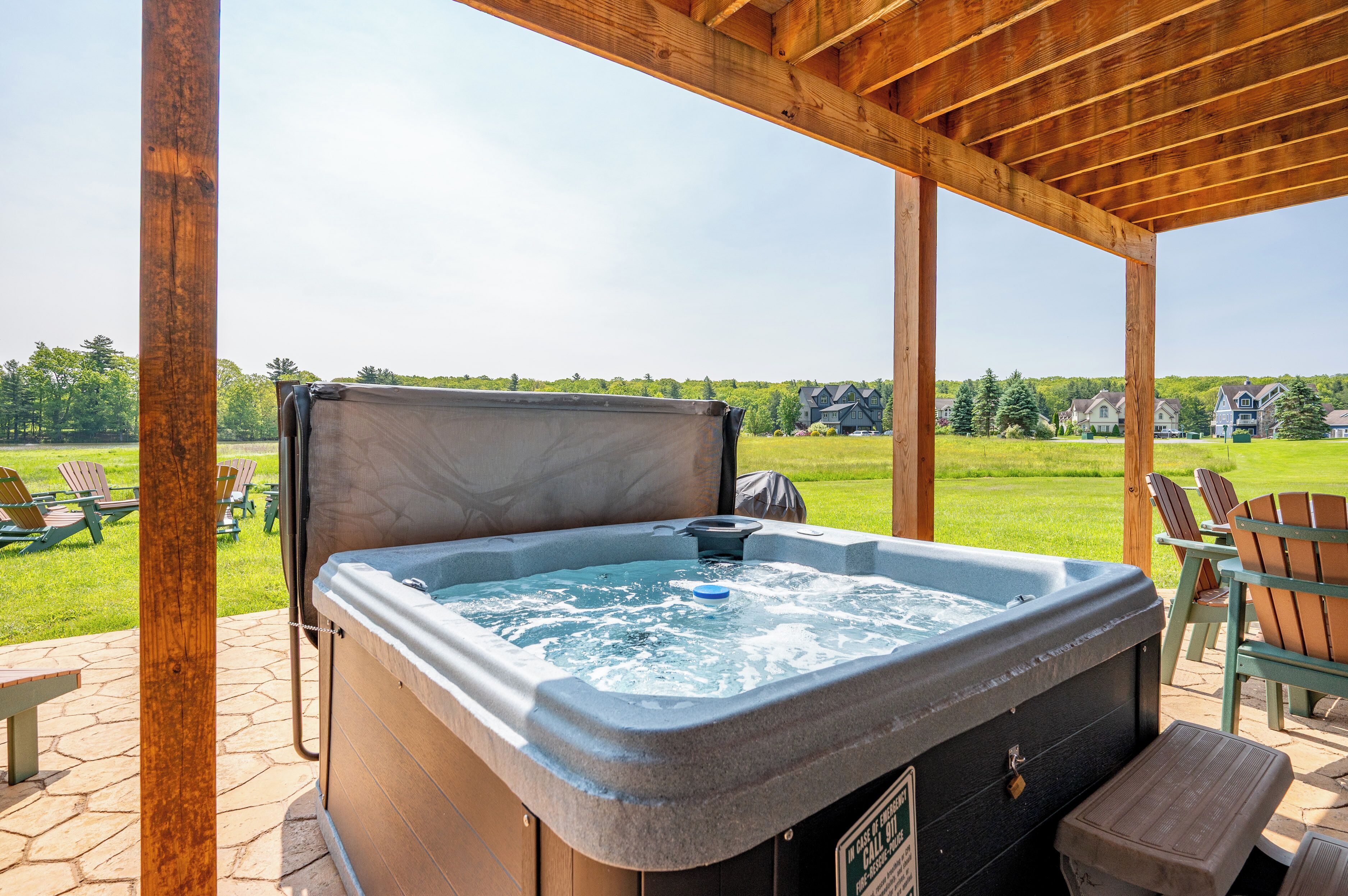 A hot tub under a wooden pergola with a view of a grassy area and houses in the background.