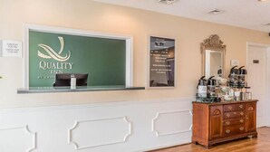 A reception area with a 'Quality Inn' sign, a check-in/out schedule, a mirror, and a coffee station. - Rodeway Inn Sylacauga Hwy 280 (Sylacauga)