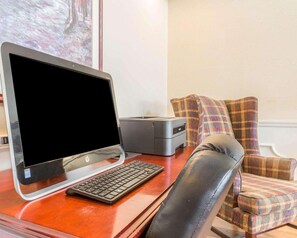 A wooden desk with a desktop computer, a keyboard, and a printer. There is a plaid-patterned chair and a painting on the wall.